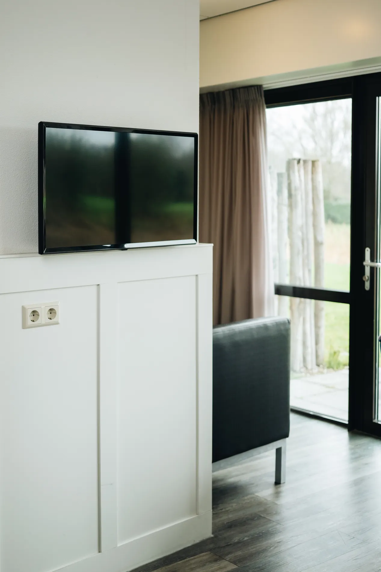 Moderne flatscreen televisie gemonteerd aan een witte wand in een lichte kamer met uitzicht op de natuurlijke omgeving.